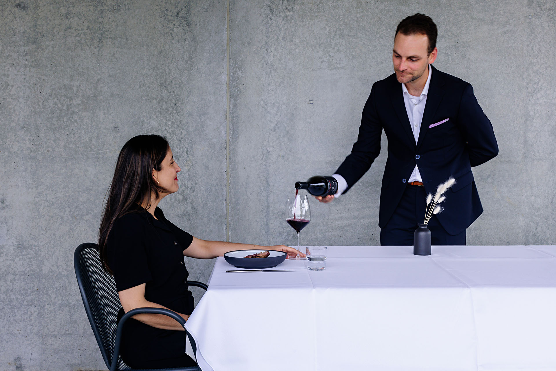 A woman sits at a dining table with a white tablecloth and a single plate, looking up as a man in a suit pours red wine into a glass. There's also a black vase with white flowers on the table, and a concrete wall in the background.