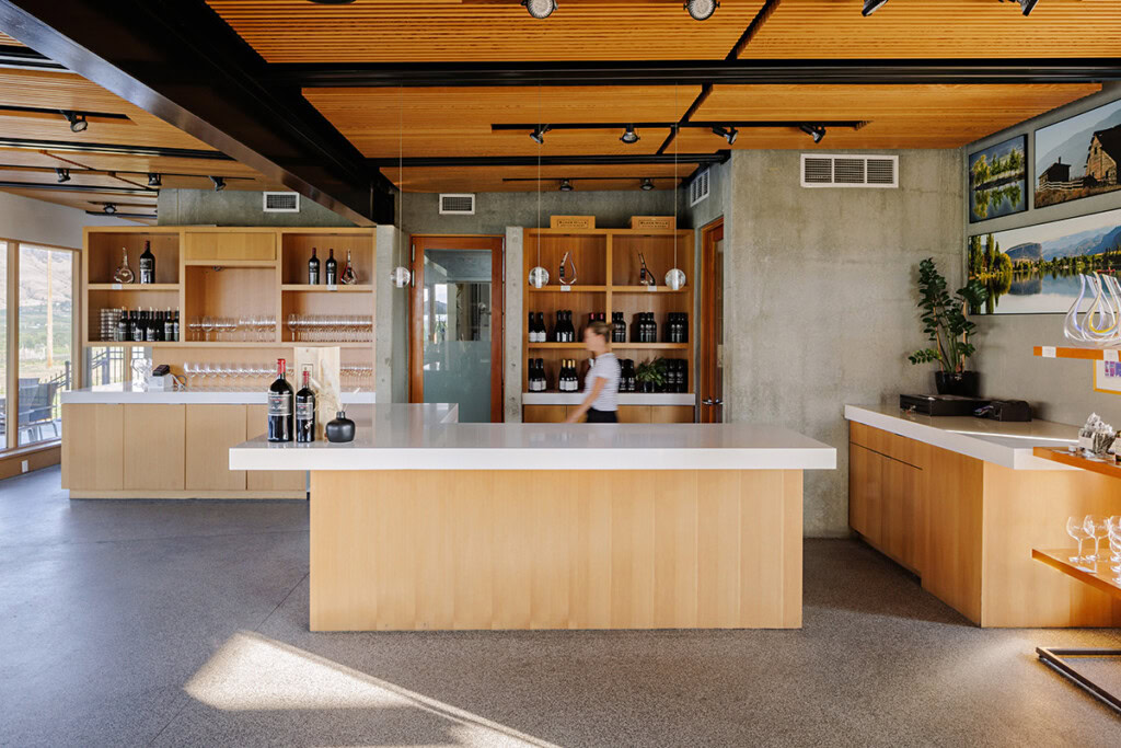 A modern wine tasting room featuring sleek wood and concrete finishes. A staff member is visible in the background, slightly blurred, moving behind the counter. The room includes shelves with wine bottles and glasses, framed artwork, and potted plants for décor.