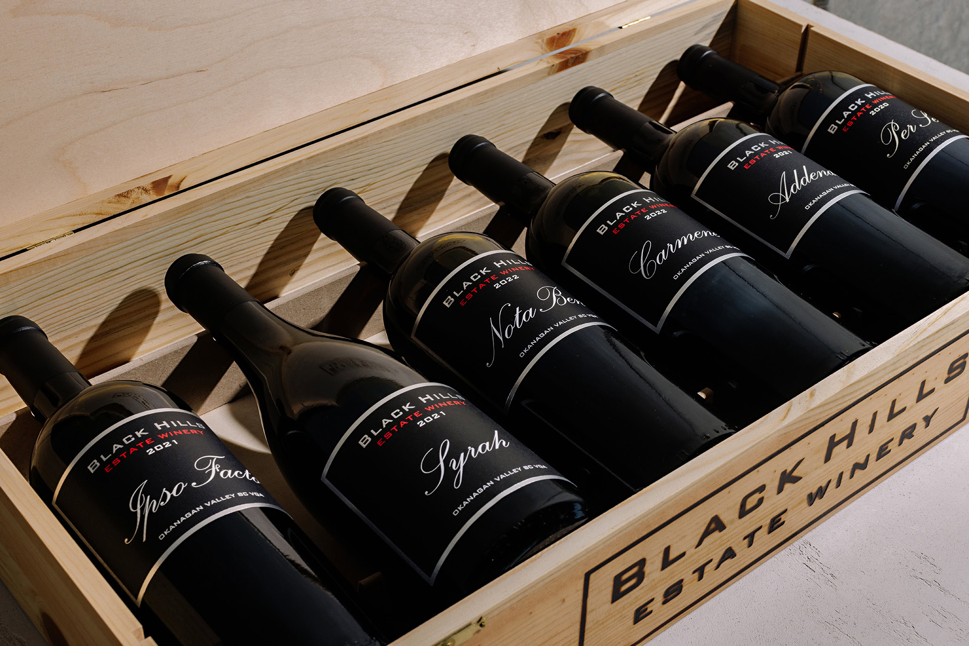 A wooden crate containing seven bottles of Black Hills Estate Winery red wine, each with a black label and distinct names like Syrah, Per Se, Nota Bene, and Carmenere. The crate lid is partially open, revealing the neatly arranged bottles inside.