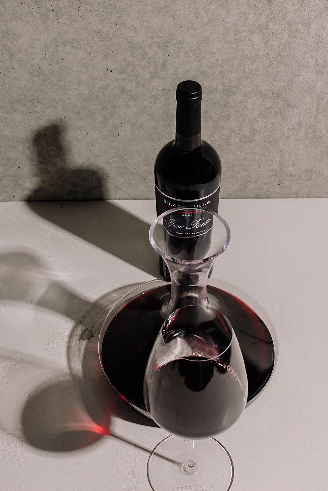 A bottle of red wine stands next to a half-filled wine decanter and a nearly full wine glass on a white surface. The bottle casts a shadow on the gray background, while the wine glass is reflected below it.