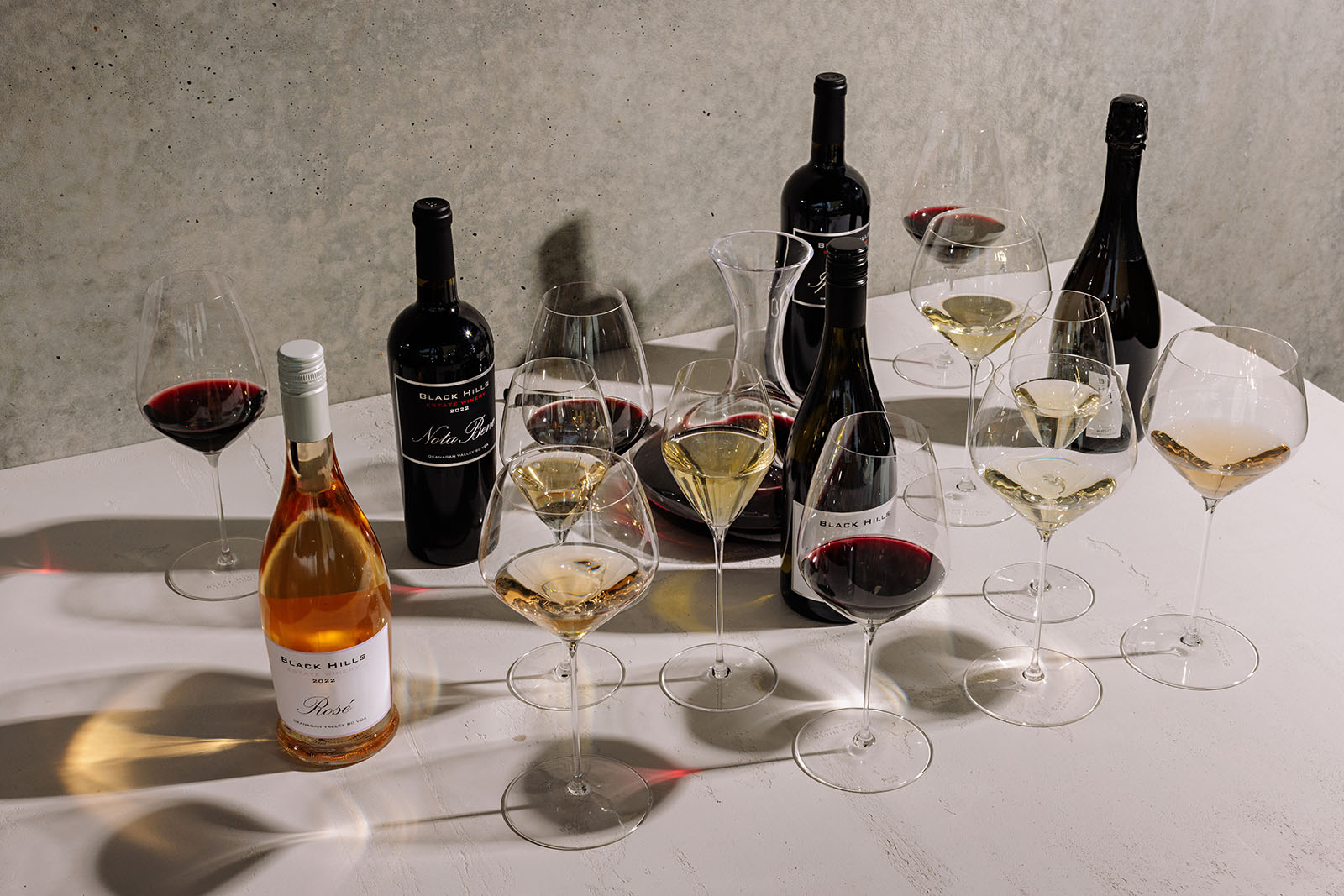 A table with an assortment of wine bottles and glasses. There are two red wine bottles and one rosé wine bottle, with corresponding glasses filled with wine. A wine decanter is surrounded by multiple glasses containing red and white wine. The background is a plain, light-colored wall.