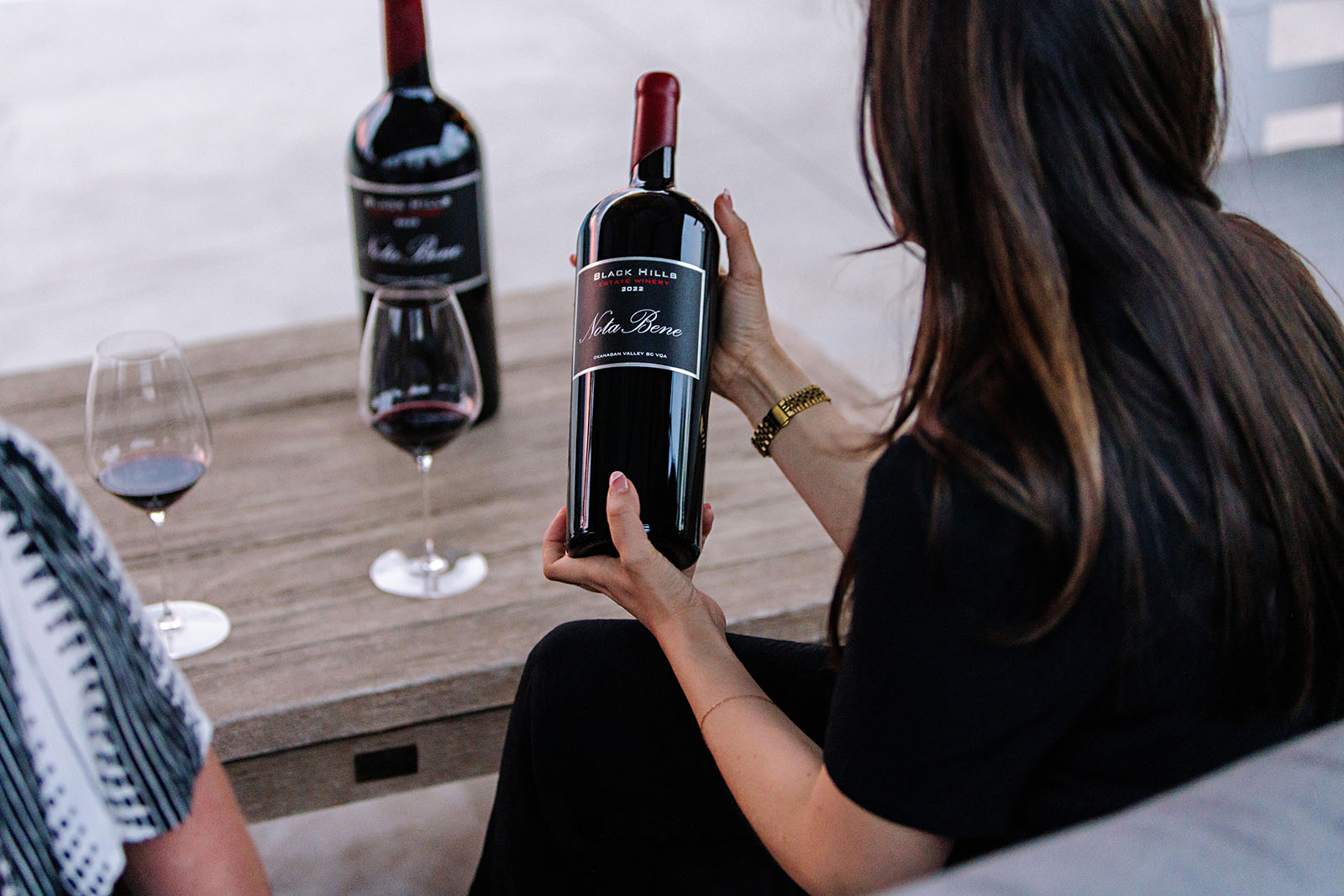 A person with long hair, dressed in black, is sitting at a wooden table holding a large bottle of red wine labeled 