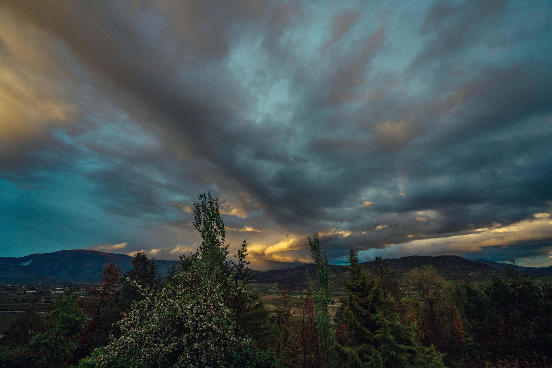 A dramatic landscape with a cloudy sky at dusk. The image shows dark, moody clouds with patches of sunlight breaking through. Below, there are lush green trees and rolling hills, with distant mountains providing a serene backdrop.