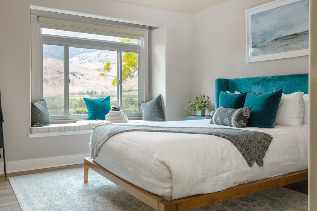 A modern bedroom featuring a large window with a scenic view, a wooden bed frame with white bedding, and teal accent pillows and headboard. A painting and plants add decoration to the room, which also has a gray rug and a stack of white towels on the window bench.