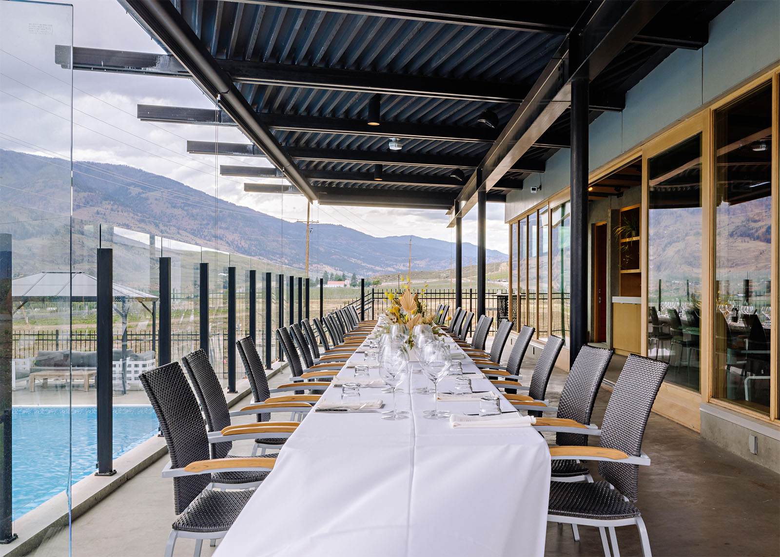 A long dining table set with white linens, glasses, and tableware is arranged under a covered patio with a view of mountains and a pool. The table is flanked by black wicker chairs and the area is enclosed by glass panels. The setting is elegant and scenic.