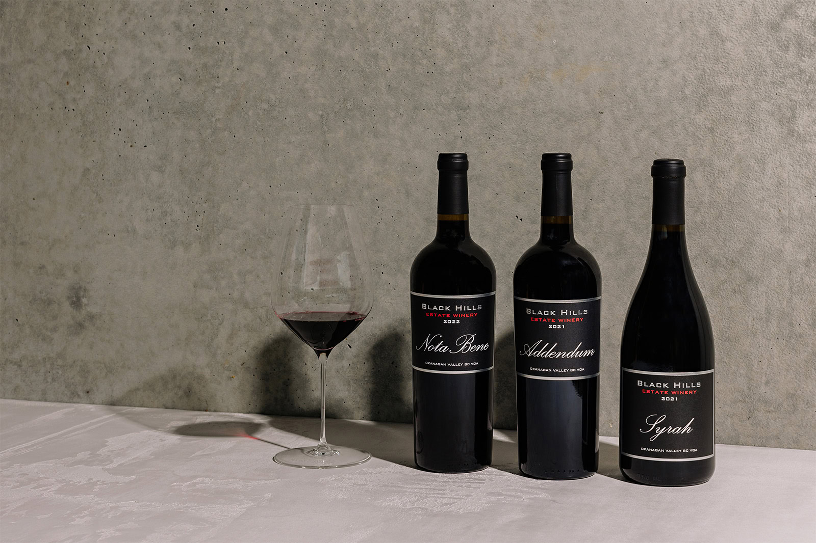 Three bottles of Black Hills Estate Winery red wine are placed on a white surface against a gray, textured background. A partially filled wine glass stands to the left of the bottles. The wine varieties are Syrah, Nota Bene, and Addendum.