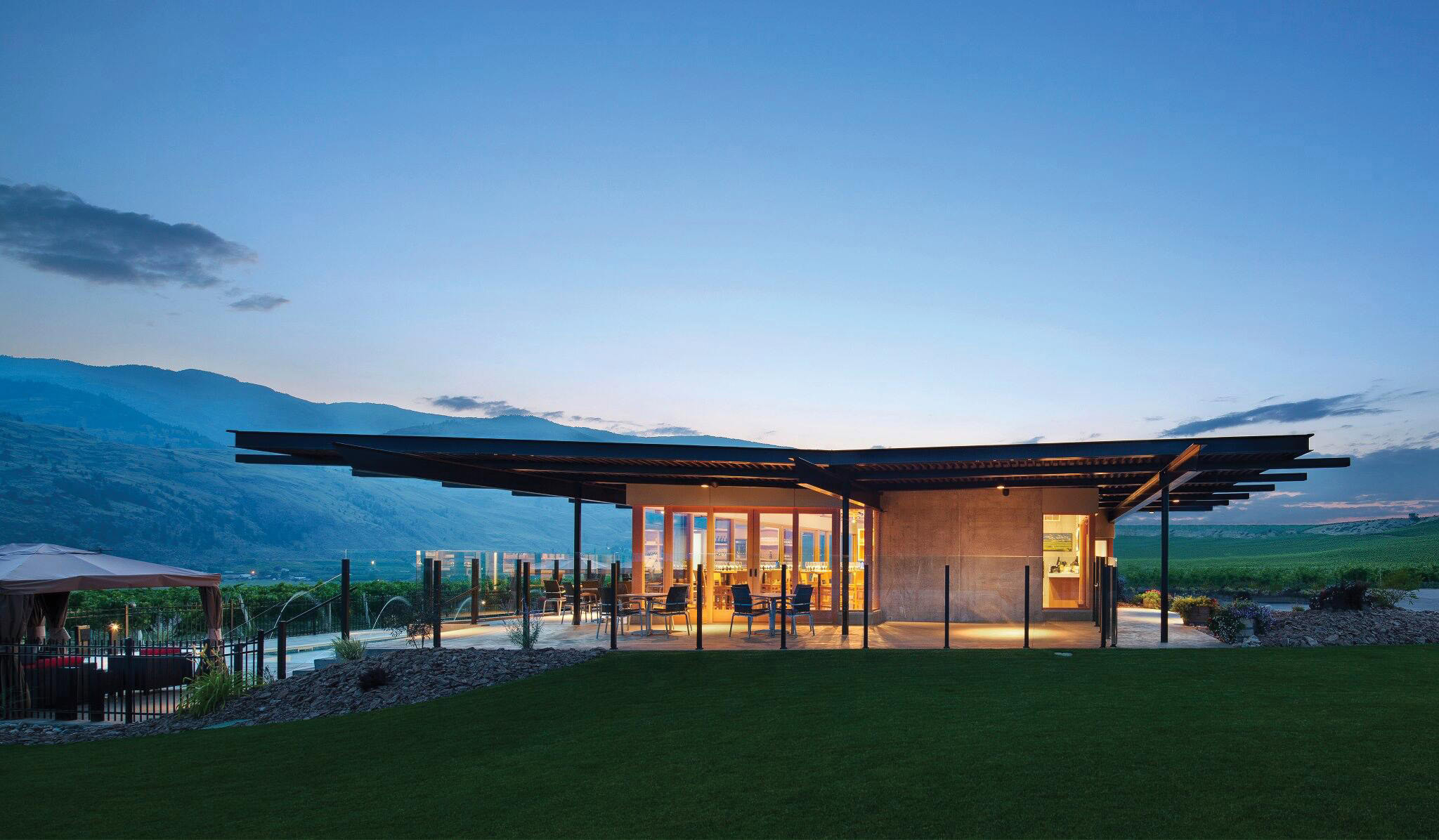 A modern building with large glass windows and an expansive roof is illuminated from within at dusk. The structure is surrounded by green lawns and a distant mountainous landscape. The sky is clear with a gradient from blue to dark blue as twilight sets in.