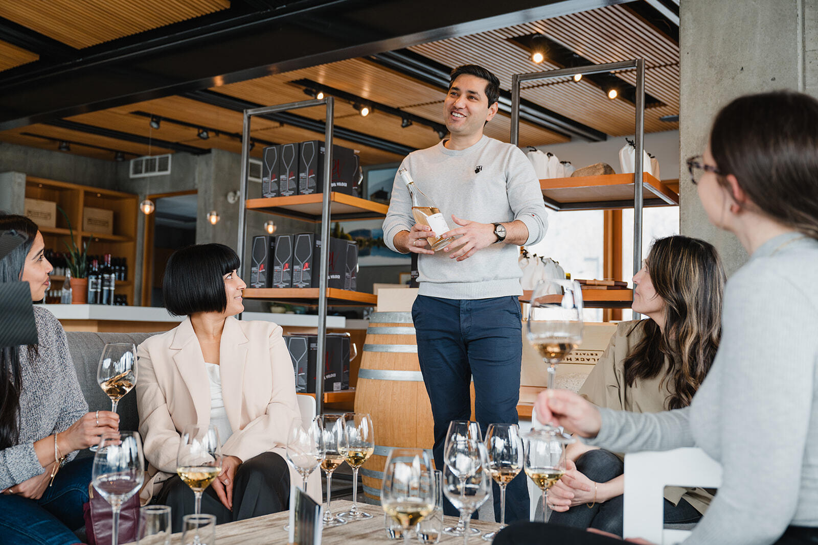 A group of people are gathered in a modern wine-tasting room. A man stands confidently holding a wine bottle and addressing the seated group, all of whom are holding wine glasses and attentively listening. The room has contemporary decor with wooden accents.