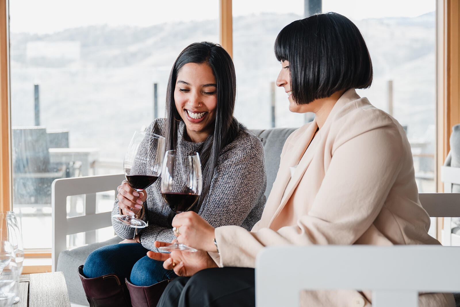 Two women are sitting and holding glasses of red wine, smiling and engaged in conversation. They are indoors, with large windows showing a blurred outdoor view of hills or mountains. One woman is wearing a sweater, and the other is in a light-colored blazer.