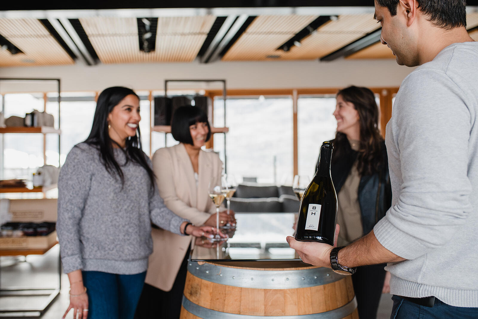 A person presents a bottle of white wine to three people standing around a wooden barrel in a modern, light-filled room. The three people appear engaged and smiling, holding glasses of wine. The room has large windows and contemporary decor.