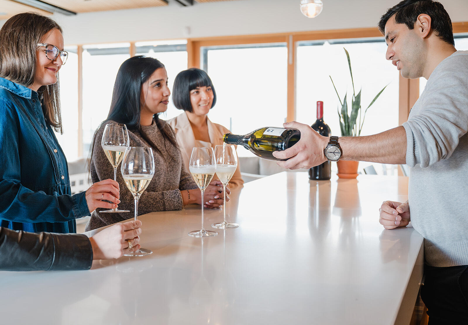 A man in a light sweater pours white wine into a glass held by a woman. Three other women stand at the counter, each holding a wine glass and watching the man pour. The background features a bright, modern interior with large windows and a plant.