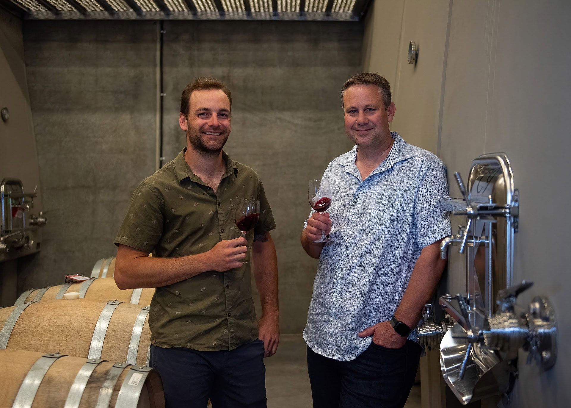 Two men stand in a winery, each holding a glass of red wine. They are smiling and appear relaxed, with one man wearing a green shirt and the other in a light blue shirt. Behind them are wooden wine barrels and stainless steel equipment against a concrete wall.