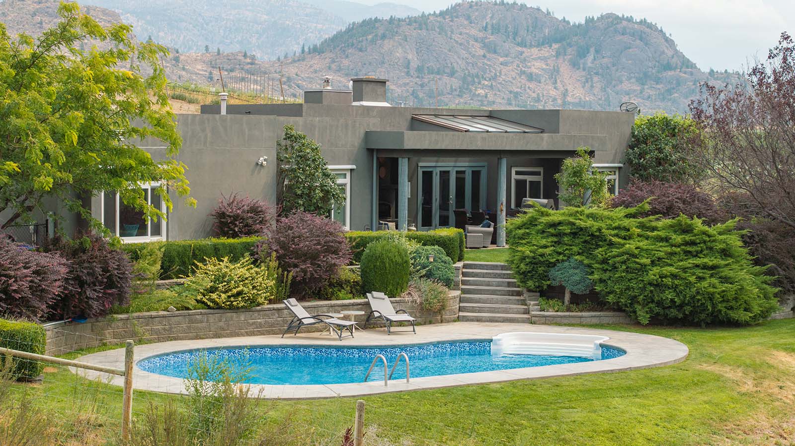 A modern house with large windows and patio is situated amidst a landscaped garden with various shrubs and trees. In front, there is an oval-shaped swimming pool surrounded by a lounge area with two pool chairs. Mountains are visible in the background.