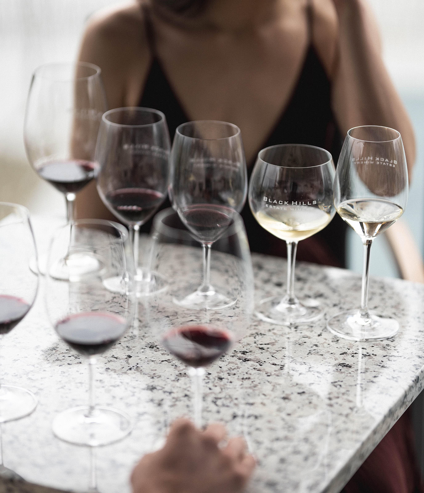 A person sitting at a table with an assortment of wine glasses, both red and white wine, on a marble surface. The person appears to be wearing a black outfit, and the atmosphere suggests a wine tasting event. Some wine glasses have the text 