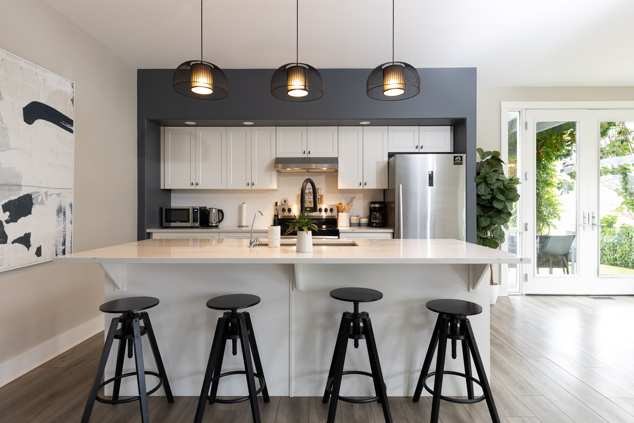 Modern kitchen featuring a large island with four black stools. Overhead, three black pendant lights illuminate the space. The kitchen has white cabinets, stainless steel appliances, and a door leading to an outdoor area with greenery visible through glass doors.