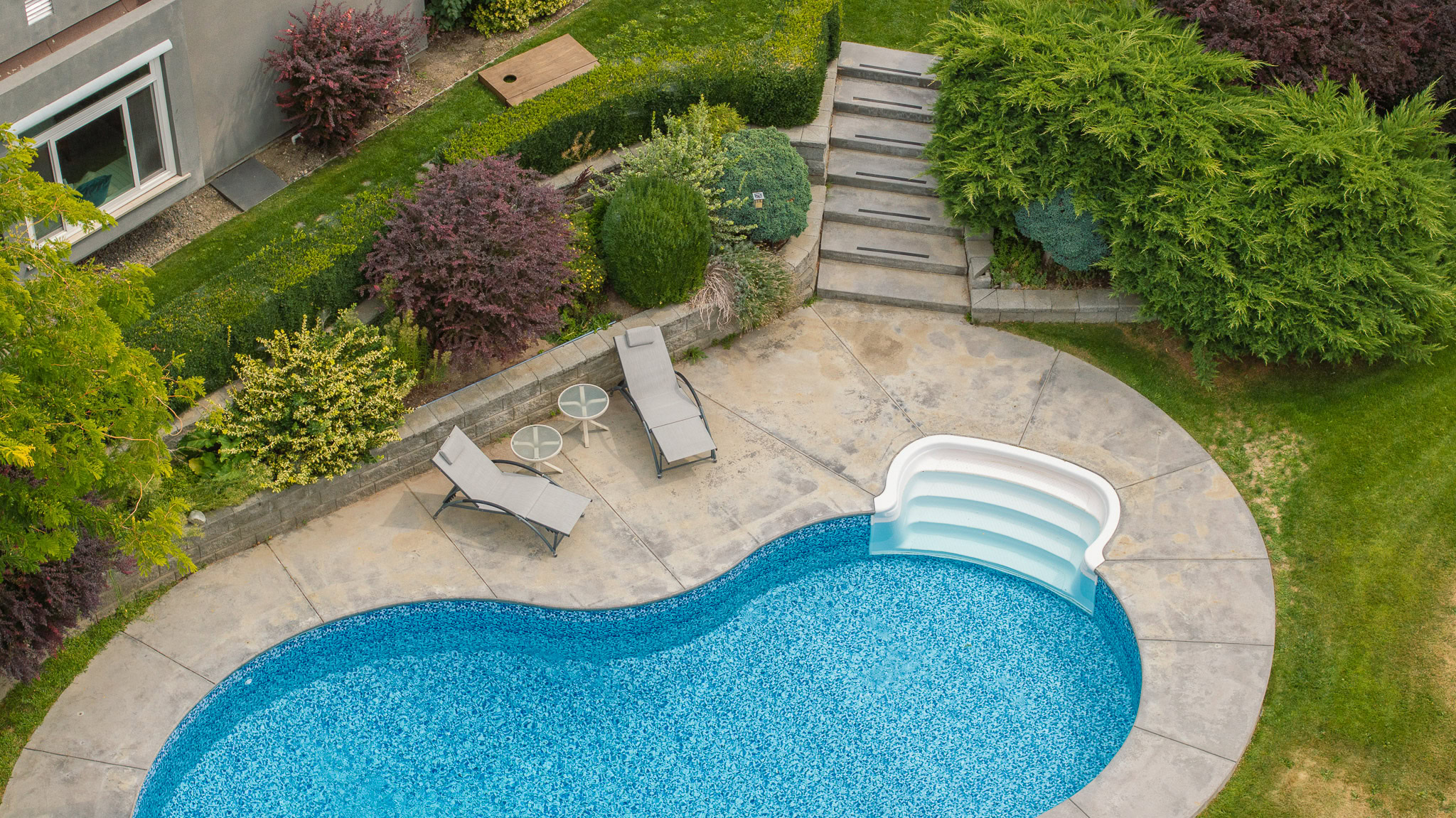 Aerial view of a backyard featuring a kidney-shaped swimming pool with a built-in staircase. The pool is surrounded by a stone patio with two lounge chairs and a small table. Lush green bushes and plants adorn the landscape, and stone steps lead to an upper level.