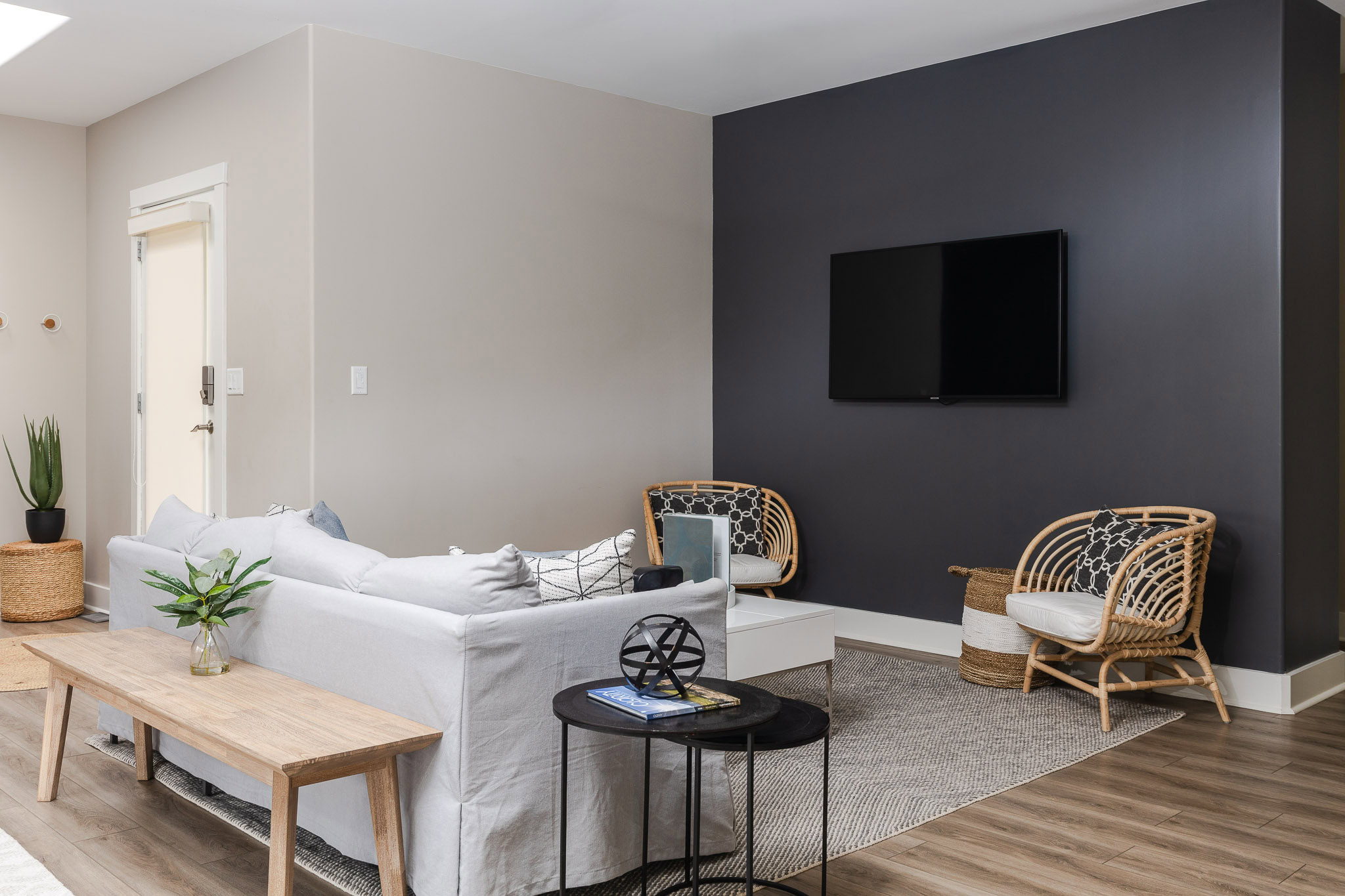 A modern living room features a sofa with white cushions, a wooden coffee table, and a TV mounted on a dark accent wall. Two wicker chairs with cushions are set on a gray rug, and a small black side table holds a decor item and a book. Wooden floors complete the look.