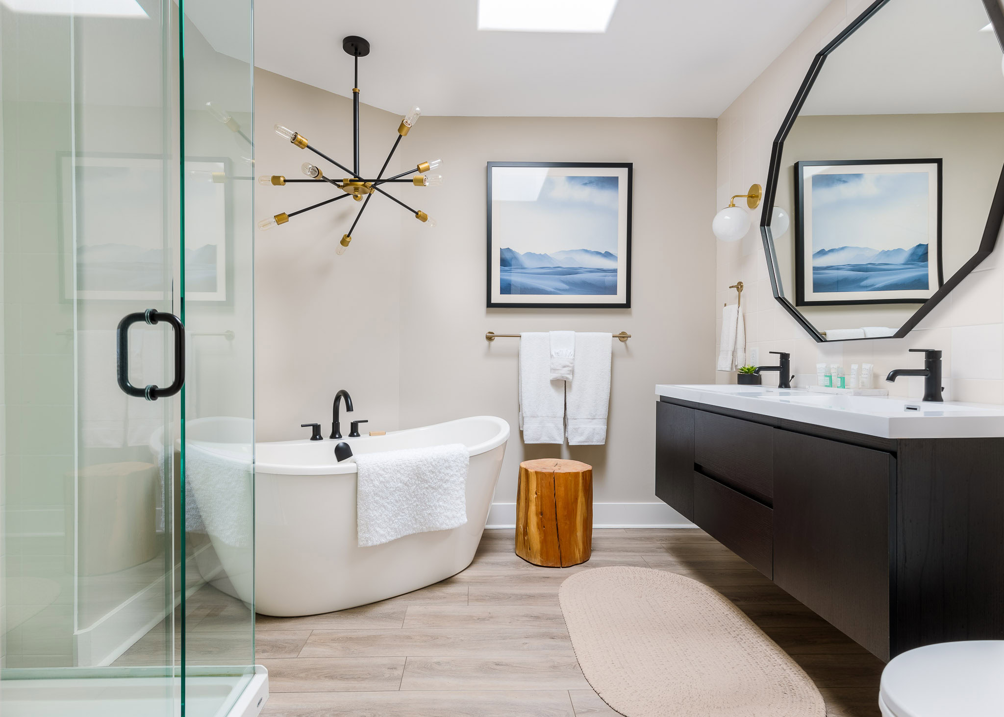 Modern bathroom with a freestanding bathtub beside a wooden stool, enclosed glass shower, and a wall-mounted dual-sink vanity with black fixtures. The room features two framed artworks, a geometric pendant light, and ample natural light from a skylight.