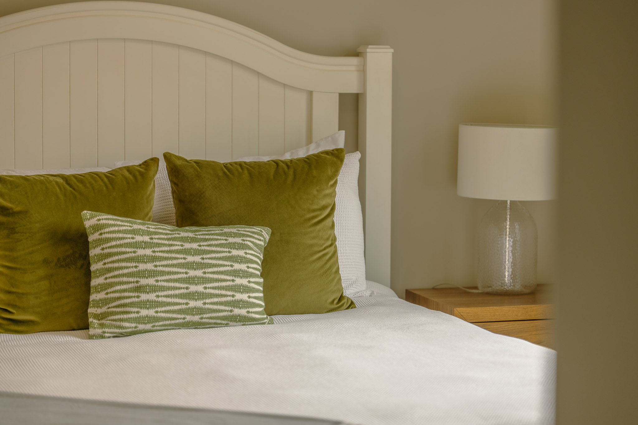 A cozy bedroom features a white bed with a white headboard and is adorned with green accent pillows. A green patterned rectangular pillow sits in front of two larger green pillows. A wooden nightstand holds a glass lamp with a white shade.