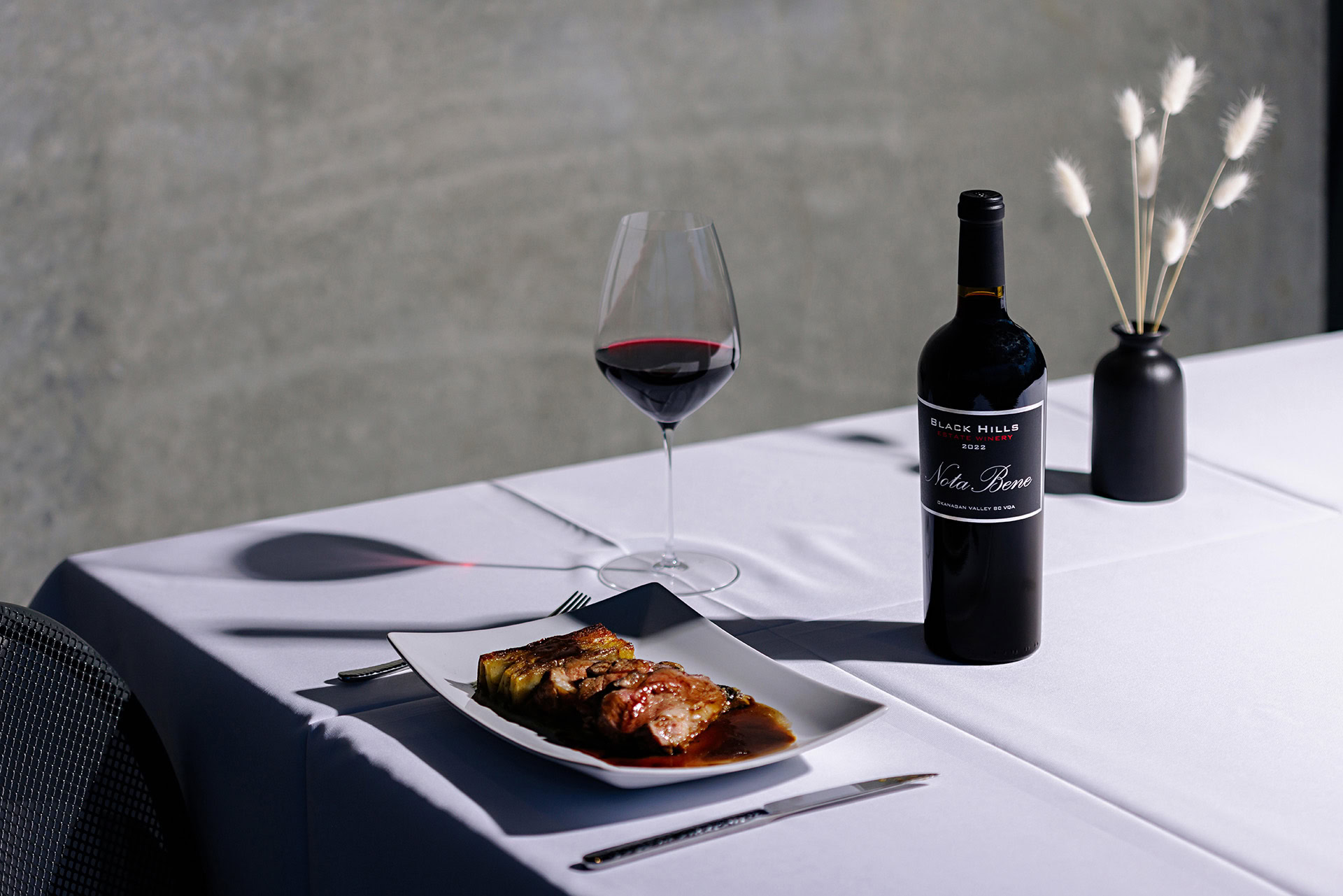 A neatly set table featuring a white tablecloth holds a dish of elegantly plated food, a bottle of red wine, a filled wine glass, a fork, and a knife. Nearby, a small black vase with dried white flowers adds a decorative touch. The background is soft and neutral.